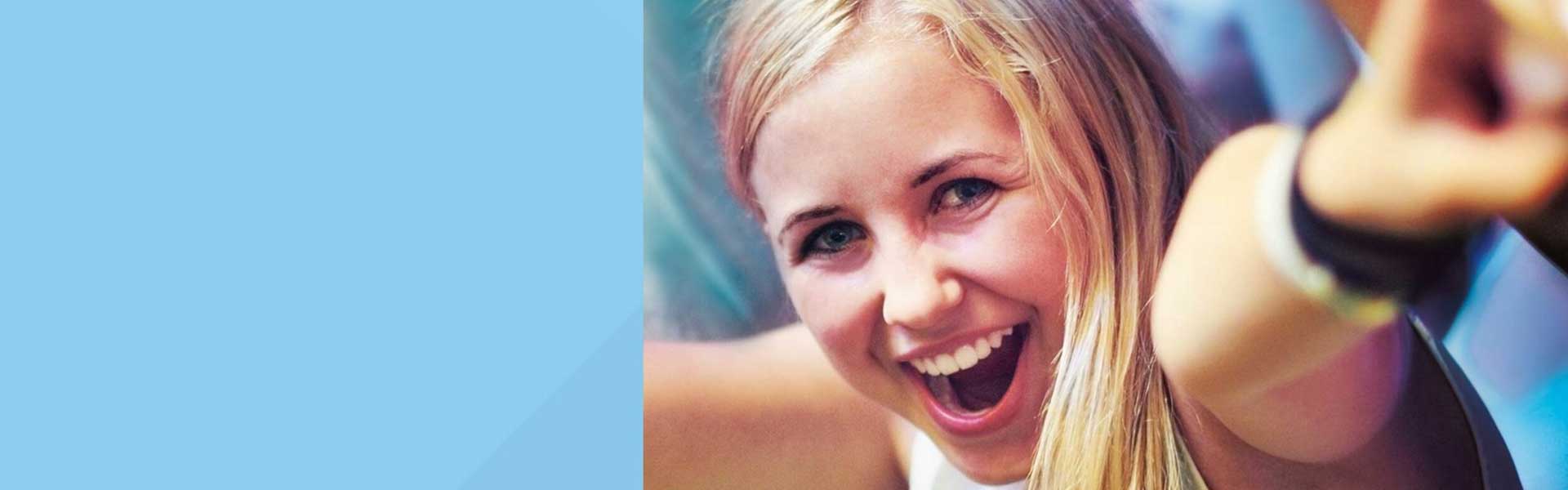 Junge, blonde Frau ruft lachend etwas direkt in die Kamera und hebt dabei die Arme